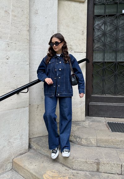 Chaqueta de mezclilla y jeans de pierna ancha en azul oscuro. La chaqueta cuenta con botones y bolsillos, con un cuello. Camiseta blanca debajo. Zapatos negros.