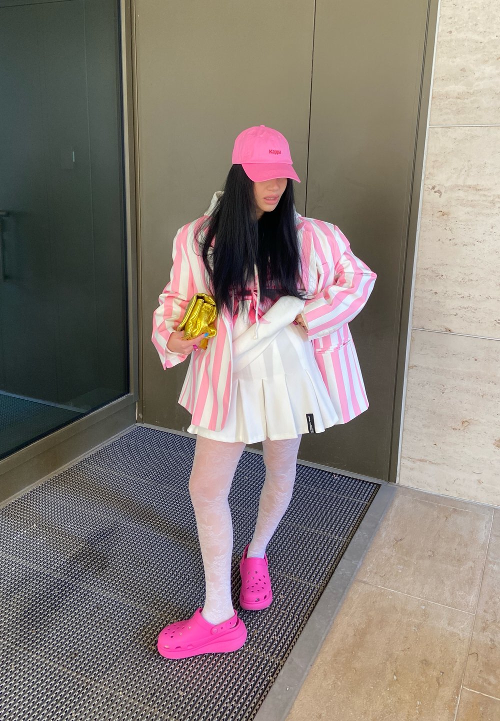 Chaqueta de rayas rosa y blanca de gran tamaño sobre una sudadera negra, combinada con una falda blanca con pliegues, medias estampadas y Crocs rosas. Bolso de mano dorado.
