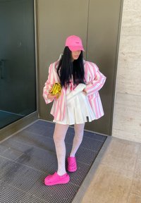 Chaqueta de rayas rosa y blanca de gran tamaño sobre una sudadera negra, combinada con una falda blanca con pliegues, medias estampadas y Crocs rosas. Bolso de mano dorado.