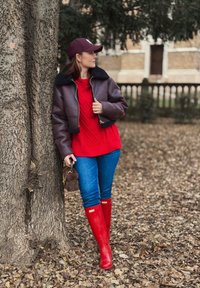 Jachetă de piele maro, scurtă, cu guler de blană neagră, pulover roșu cu model împletit, blugi albaștri și cizme strălucitoare roșii din cauciuc. Ține o geantă de blană maro.