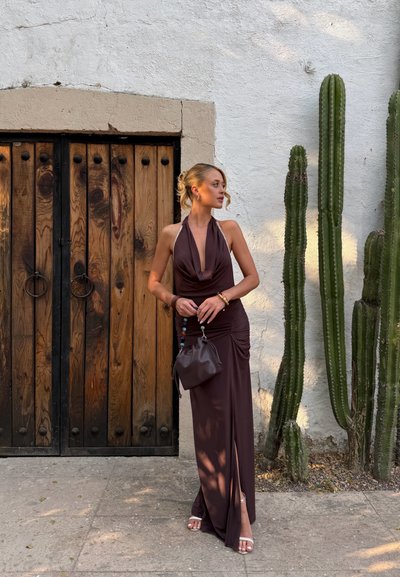 Mujer con vestido marrón sin mangas que sostiene un pequeño bolso, de pie junto a puertas de madera y altos cactus contra una pared blanca texturizada.
