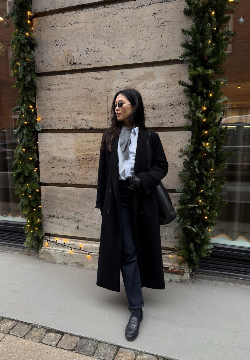 Long manteau noir sur une chemise bleu clair, jean foncé et mocassins noirs. Debout près d'un mur en pierre texturé avec des guirlandes vertes et des lumières.