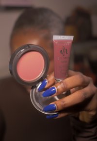 Rund, mat blush-kompakt med pink pulver; lille tube med flydende mat makeup i en afdæmpet pink nuance, holdt af en hånd med blå negle.