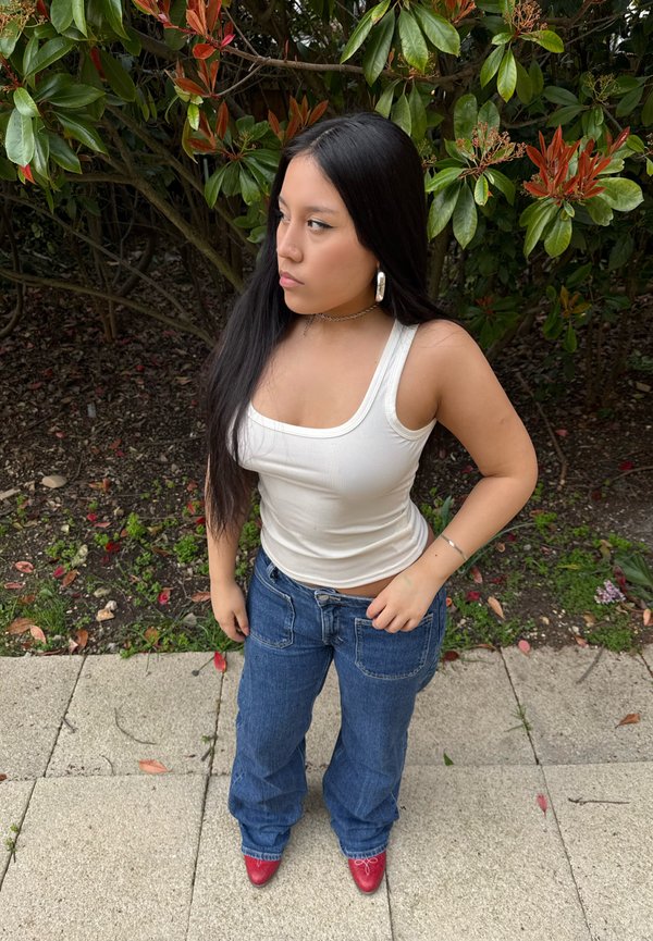 Top de tirantes blanco con canalé, jeans amplios azules y zapatos de tacón rojos. Cabello largo y oscuro, pendientes de aro plateados, de pie contra un fondo de vegetación y pavimento.
