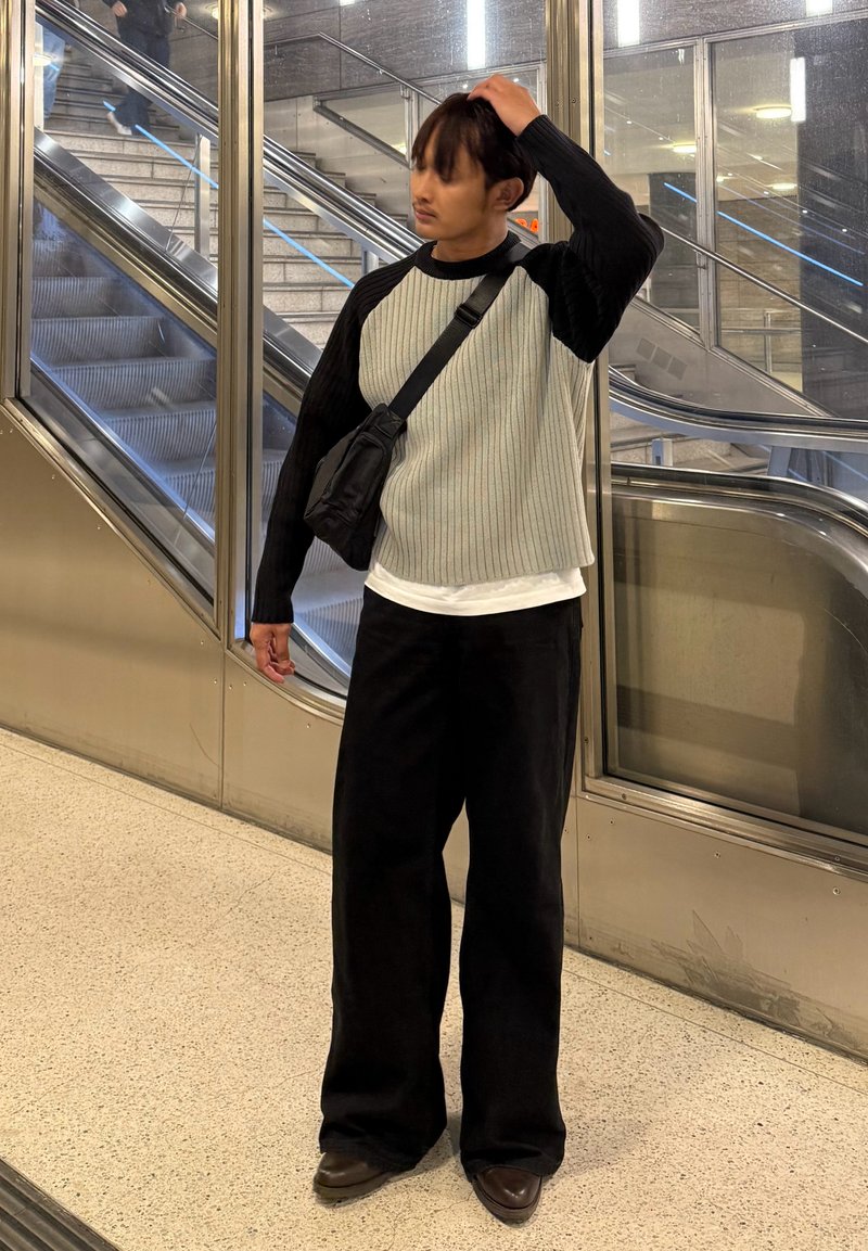 Une personne se tient à l'intérieur portant un pull côtelé noir et gris, un pantalon noir à jambes larges et des chaussures marron, avec un petit sac noir en bandoulière.