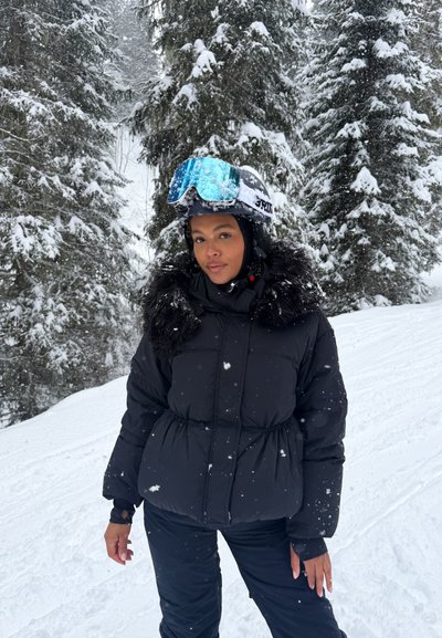 Persona con chaqueta de invierno negra con cuello de piel y casco de ski con gafas azules, de pie en un bosque nevado con la nieve cayendo.