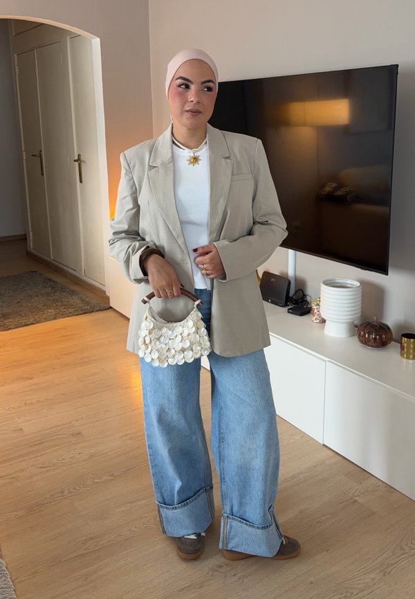 Mujer con blazer beige y vaqueros azul claro sostiene un bolso blanco adornado con conchas en un salón con televisor montado y elementos decorativos.