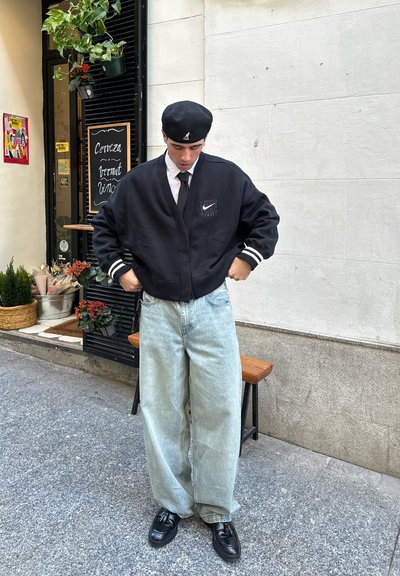 Chaqueta bomber negra de Nike con rayas blancas, jeans holgados azul claro y zapatos negros. Plantas en macetas en el fondo, pared de piedra.