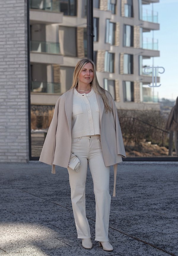 Žena stojaca vonku v béžovom kabáte cez biely sveter a nohavice, drží malú bielu tašku, za ňou je moderná budova s odrazom v sklenených oknách.