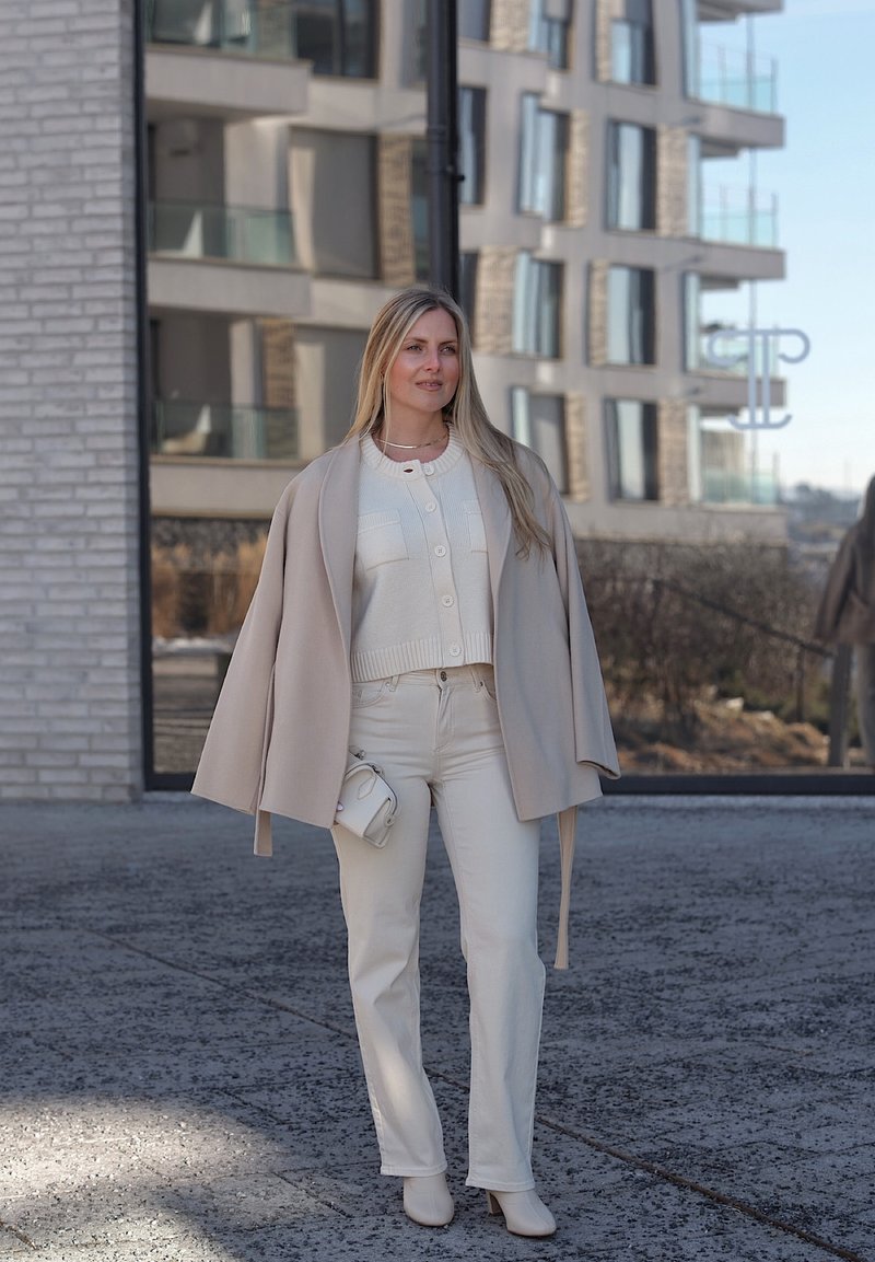 Frau steht im Freien in einem beigen Mantel über weißer Strickjacke und Hose, hält eine kleine weiße Tasche, mit modernem Gebäude und Spiegelung im Glasfenster im Hintergrund.