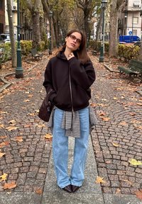 Juodas užsegamas džemperis su užtrauktuku, languota marškinių sluoksnis, šviesiai mėlynos plačios kelnės ir ruda rankinė; stovintis ant akmenų grindinio su rudens lapais.