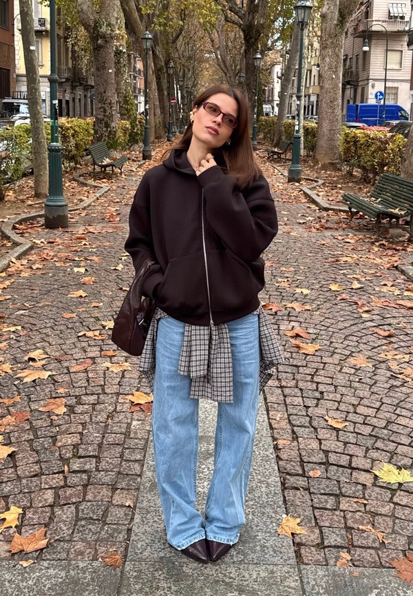 Sweatshirt preta com fecho de correr, camisa de xadrez por baixo, jeans azul claro de perna larga e mala castanha; a pé numa calçada de pedras com folhas de outono.
