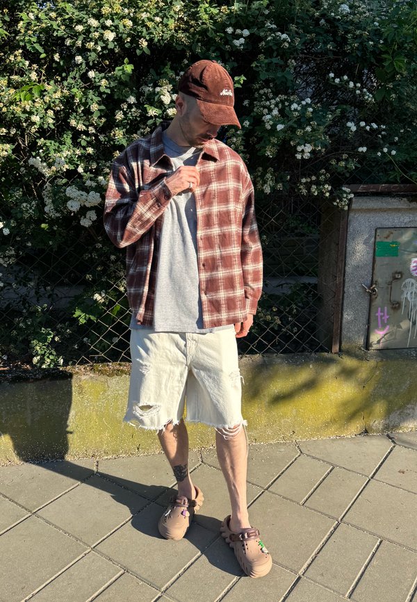Mann mit brauner Mütze, kariertem Hemd, grauem T-Shirt, zerrissenen weißen Shorts und beigen Clogs steht auf einem gepflasterten Gehweg nahe eines Zauns mit weißen Blumen.