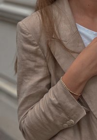 Μπεζ λινό σακάκι με υφή, συνδυασμένο με ένα απλό λευκό πουκάμισο. Χρυσό στρογγυλό βραχιόλι κοσμεί τον καρπό.