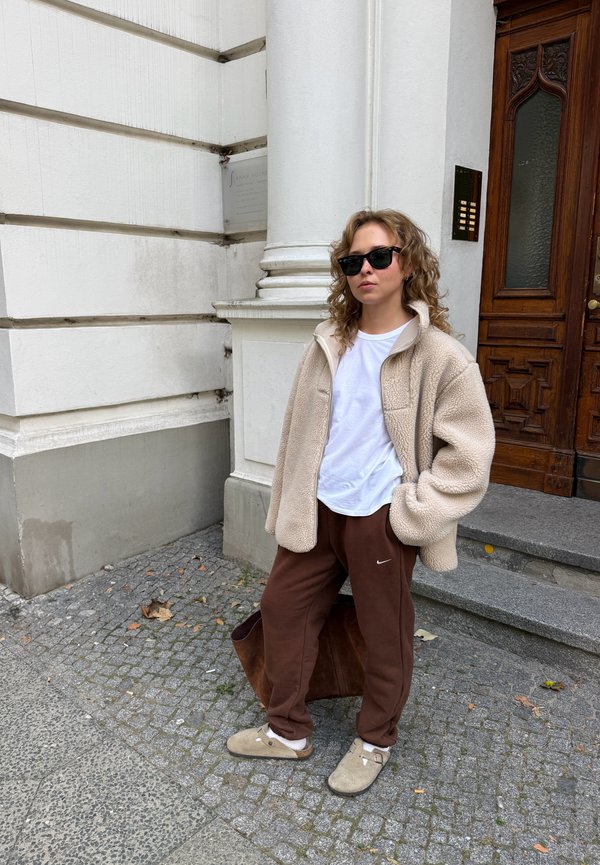 Una persona con gafas de sol, una chaqueta polar beige, pantalones marrones y zuecos está de pie al aire libre junto a un edificio blanco y una puerta de madera.