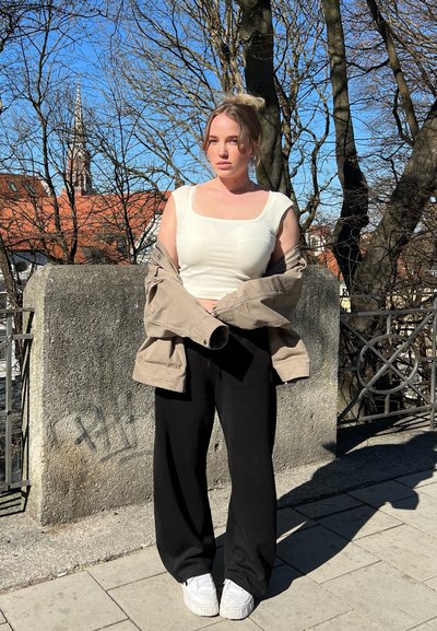 Top de tirantes blanco ajustado, chaqueta beige oversized, pantalones negros sueltos y zapatillas blancas. El fondo presenta árboles desnudos y edificios.