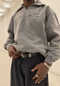 Grauer Fleece-Pullover mit Reißverschlusskragen und Logo-Patch. Schwarzer Ledergürtel mit metallischer Schnalle, Nadelstreifenhose und Ringe.