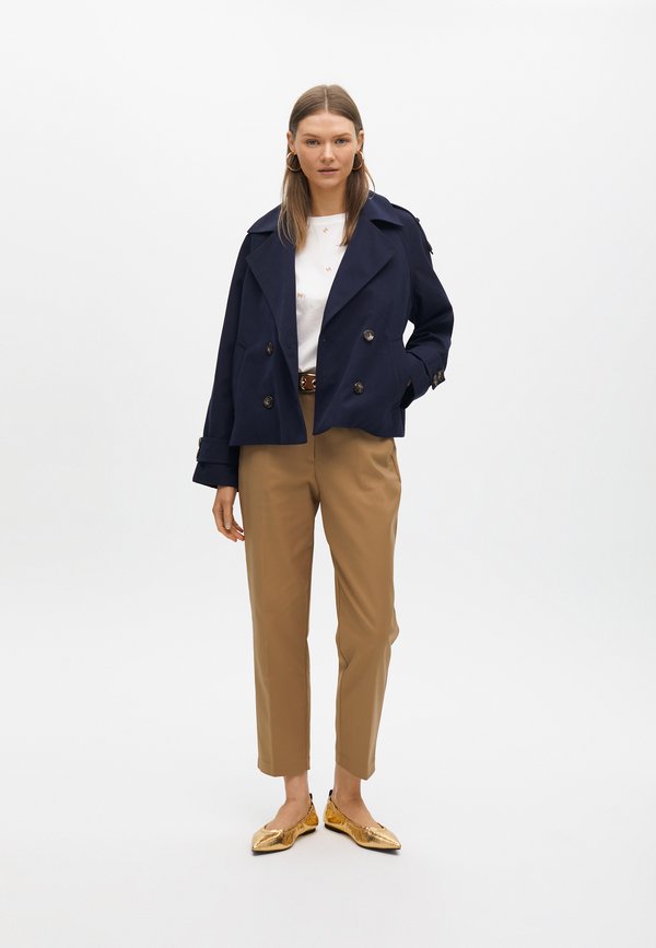 Cappotto trench navy a doppio petto, pantaloni beige e scarpe flat dorate. Il cappotto ha grandi bottoni e un colletto oversize; i pantaloni sono su misura.