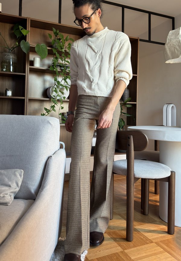 Uomo con occhiali che indossa un maglione bianco a trecce e pantaloni marroni a quadri, in piedi vicino a un tavolo da pranzo e a una mensola in legno con piante.