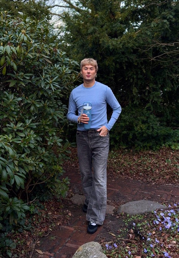 Giovane uomo con maglione blu e jeans grigi tiene una bottiglia d'acqua blu mentre cammina su un sentiero del giardino circondato da verde e fiori.