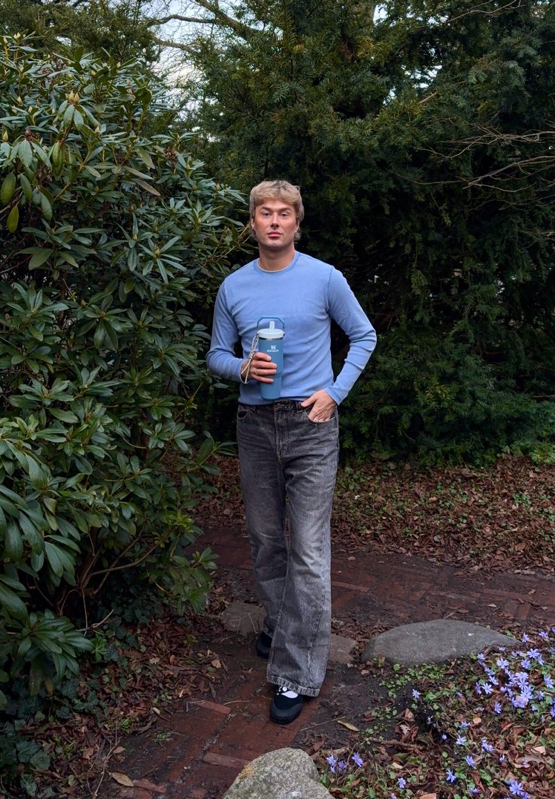 Jeune homme portant un pull bleu et un jean gris tient une bouteille d'eau bleue en marchant sur un chemin de jardin entouré de verdure et de fleurs.