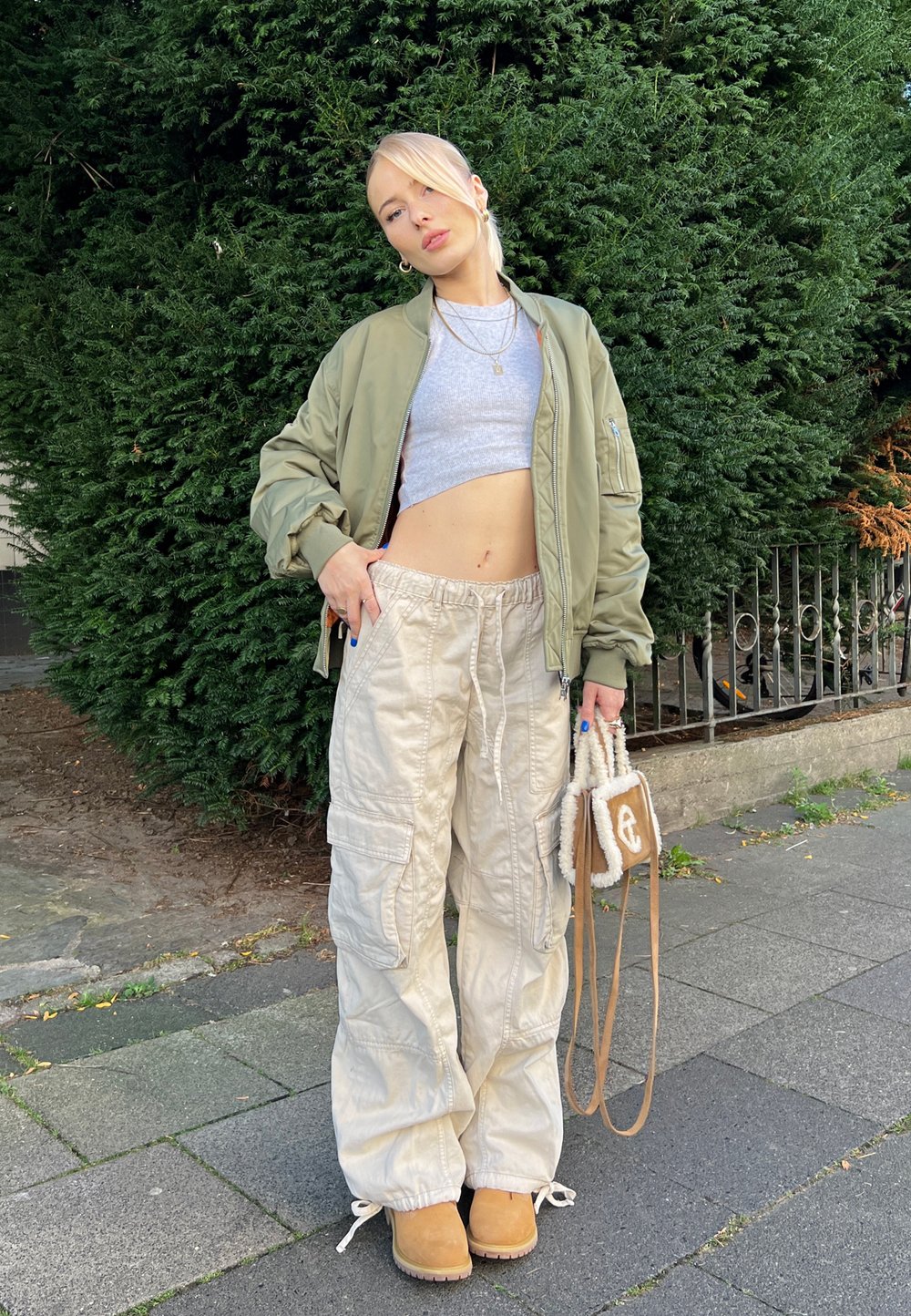 Chaqueta bomber oliva, top corto gris, pantalones cargo beige con bolsillos, botas color caña y un pequeño bolso marrón con acabado texturizado.