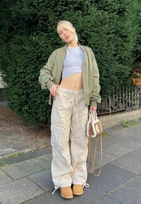 Chaqueta bomber oliva, top corto gris, pantalones cargo beige con bolsillos, botas color caña y un pequeño bolso marrón con acabado texturizado.