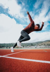 Athlète portant un haut à manches longues bordeaux et un pantalon noir, sprinte sur une piste rouge, chaussé de chaussures de sport blanches, avec un ciel bleu en arrière-plan.