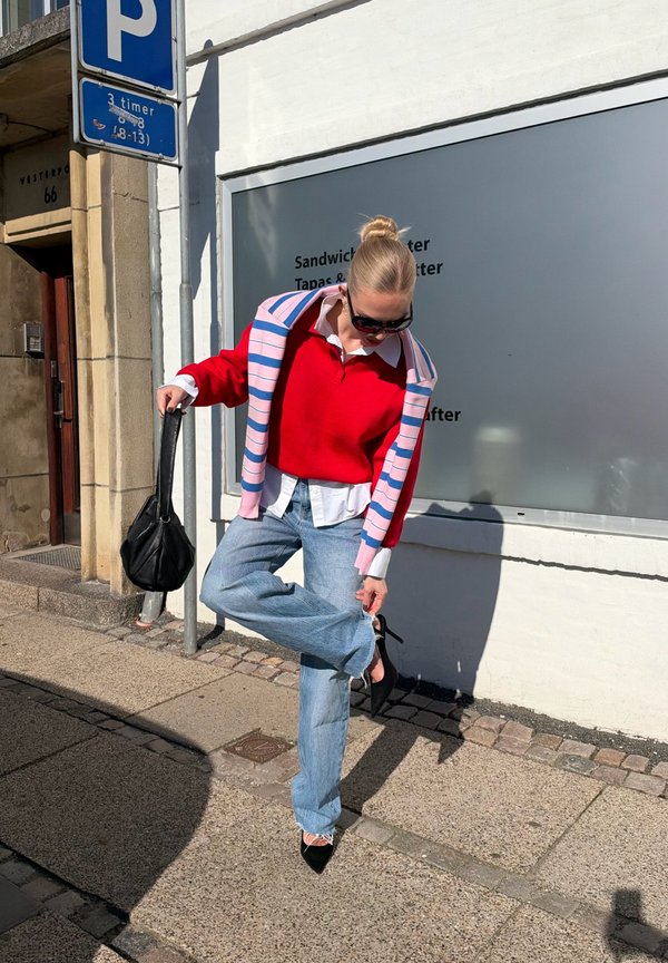 Une femme portant des lunettes de soleil tient en équilibre sur un pied tout en ajustant sa chaussure sur un trottoir ensoleillé en ville, avec un sac à main noir à la main.