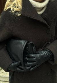 Melnas ādas somiņa, ko tur cimdiņās tvertas rokas, slāņotas zem tumša vilnas mēteļa. Ap kaklu daļēji redzams mīksts bēša šalle.