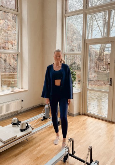 Mujer con ropa de entrenamiento oscura sosteniendo una botella de agua en una habitación luminosa con grandes ventanas y máquinas de reformador de Pilates.