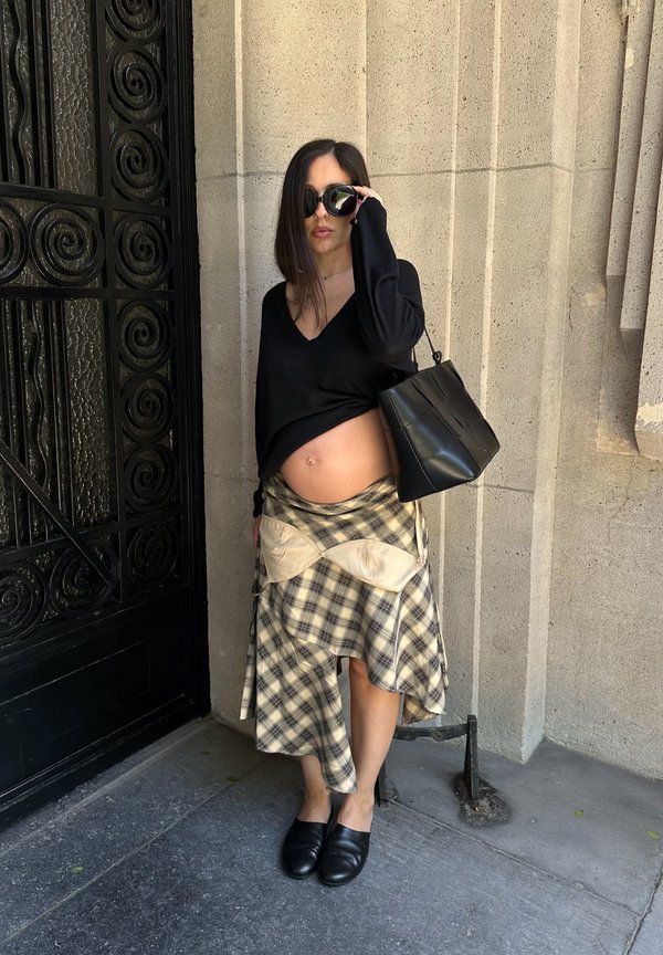 Femme enceinte portant des lunettes de soleil, un haut noir relevé pour montrer son ventre, une jupe à carreaux, des chaussures noires et un sac à main noir près d'un mur en pierre.