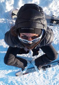 Person iført sort vinterudstyr og hvide skibriller, siddende på sne med ski krydset, kigger op mod kameraet.