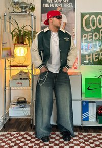 Grün- und cremefarbene Collegejacke mit gestreiften Bündchen, weißes T-Shirt, lässige blaue Jeans, silberne Kette und schwarze Sneaker mit glänzender Oberfläche.