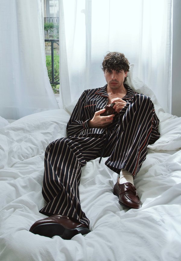 Młody mężczyzna w pasiastych piżamach i brązowych butach leży na białym łóżku, trzymając małą ciemną butelkę, za oknem przezroczyste zasłony i zieleń.