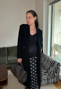 Fekete hosszú kabát fekete felső fölött, pöttyös szoknyával párosítva. Sötét textúrájú kézitáskát tart, csíkos kárpitozású szobában áll.