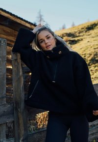 Czarny polarowy pulower z wysokim kołnierzem, przednią kieszenią na zamek błyskawiczny i dwoma złotymi guzikami jako ozdobą; przytulna, teksturowana tkanina o luźnym kroju.