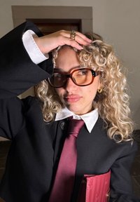 Czarna oversize'owa kurtka, biały kołnierzyk koszuli, bordowy krawat, okrągłe brązowe okulary, kręcone blond włosy, trzyma różową torebkę, z złotym pierścionkiem.