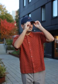 Mand iført rustfarvet striktrøje, der justerer marineblå kasket, stående på fortov i bymiljø med efterårstræer og moderne bygning i baggrunden.