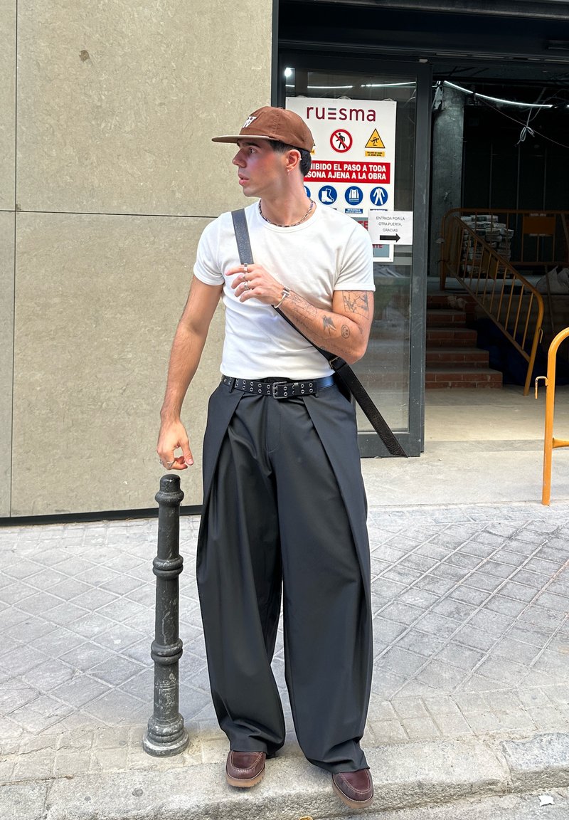 Jonge man met een bruine pet, wit t-shirt, brede zwarte broek en bruine schoenen staat op een stoep nabij de ingang van een bouwplaats.