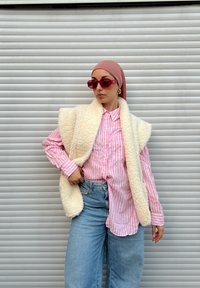 Colete branco felpudo sobre uma camisa de botão rosa às riscas, combinado com jeans azuis claros de perna larga e óculos de sol vermelhos. Fundo simples.