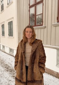 Femme aux cheveux châtain clair portant un long manteau en fourrure marron et un haut beige, debout à l'extérieur près d'un bâtiment blanc, avec de la neige au sol.