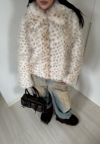 Veste en fausse fourrure à taches beiges et brunes, manches larges, associée à un jean bleu clair déchiré et un sac à main en cuir noir. Chaussures fourrées marron.