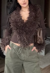 Pulover maro scurt cu guler și manșete pufoase, asortat cu pantaloni largi de culoare verde-olive. Accesorizat cu o geantă mică maro.