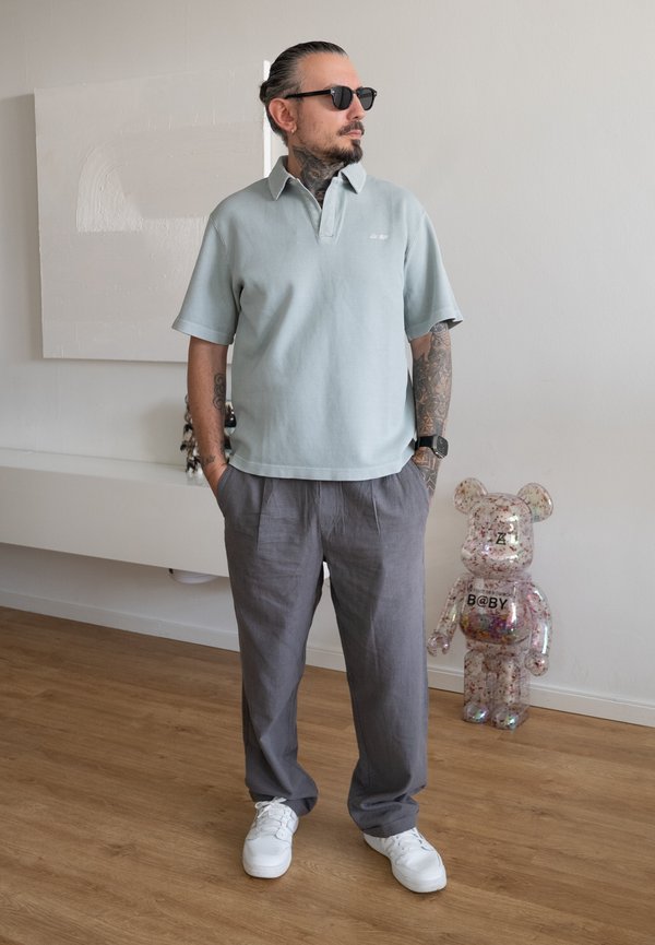Homme avec des tatouages et des lunettes de soleil portant une chemise bleu clair et un pantalon gris se tient à l'intérieur près d'une figurine d'ours en peluche transparente sur un sol en bois.