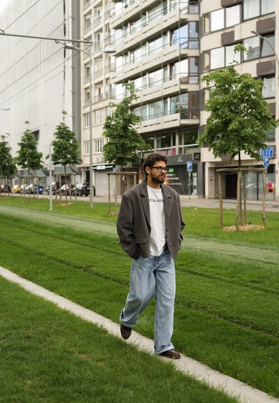 Blazer gris de gran tamaño, camiseta blanca y vaqueros azules sueltos. Caminando sobre un área con hierba, con árboles y edificios modernos al fondo.