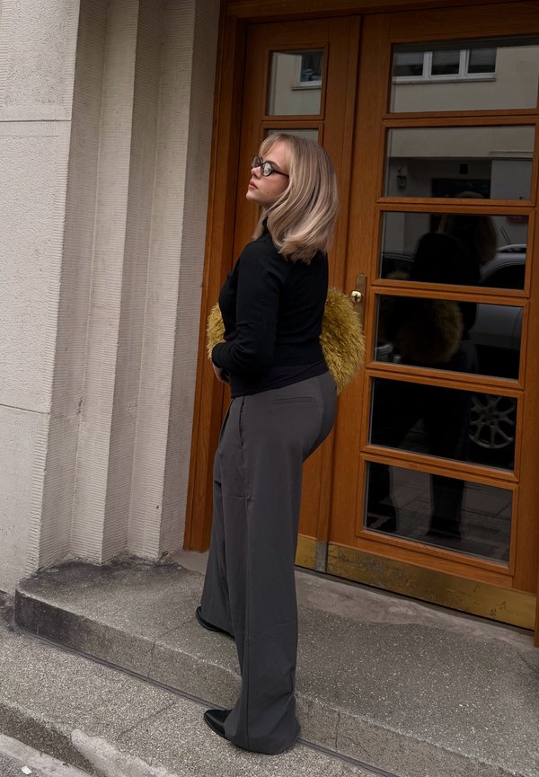 Femme aux cheveux blonds et lunettes, debout sur des marches en pierre devant une porte en bois, tenant un sac jaune pelucheux, regardant sur le côté.