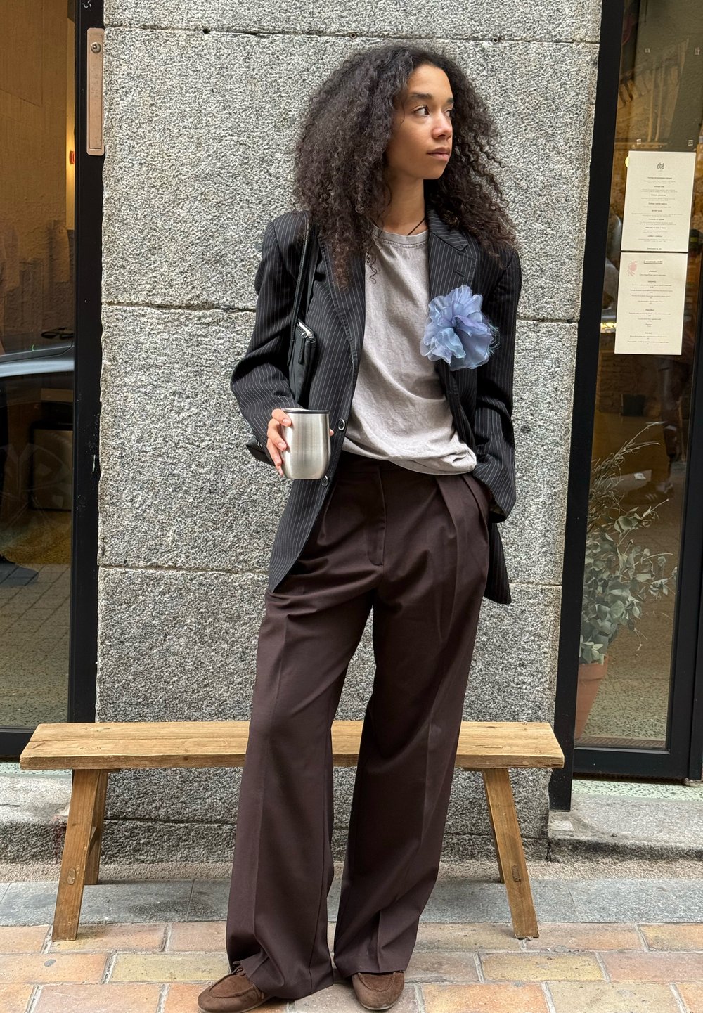 Sort baggy blazer over en grå t-shirt, brune vide bukser, blå chiffon blomst accent, sølv metal kop, og brune sko. Træbænk er synlig.