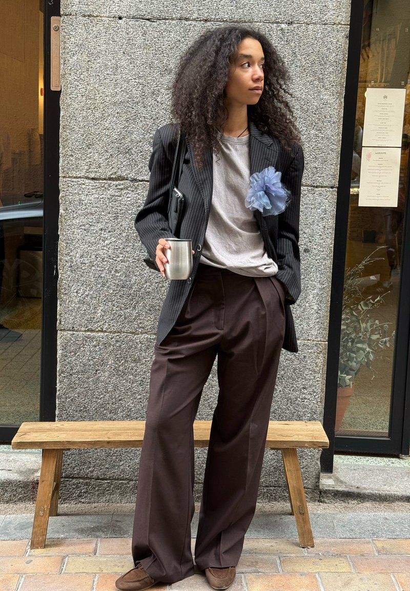 Blazer negro de rayas sobre una camiseta gris, pantalones anchos marrones, acento floral de gasa azul, taza de metal plateada y zapatos marrones. Banco de madera visible.