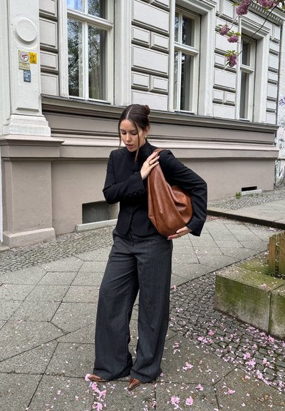 Bolso hobo de cuero marrón, blazer negro entallado y pantalones de pierna ancha a rayas grises. Pétalos de flores rosas en el suelo de adoquines.